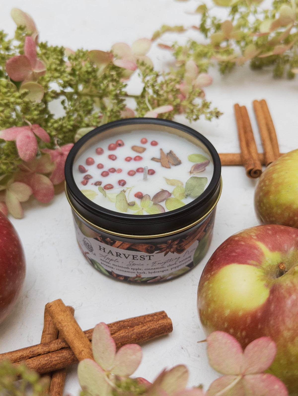 HARVEST candle tin topped with pink peppercorns and dried flowers, beside apples