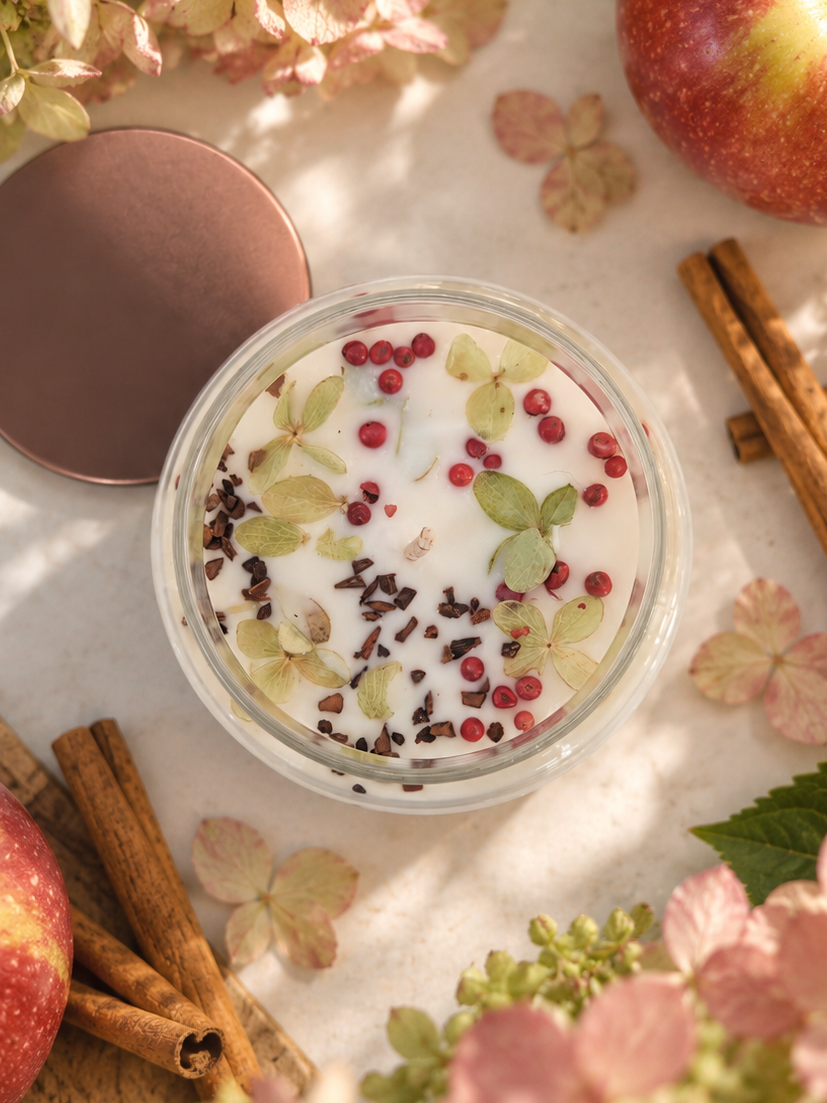 HARVEST candle in its apothecary jar, topped with pink peppercorns and dried hydrangea, beside apples and cinnamon sticks